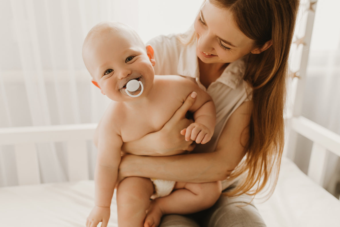 an 11 month old baby with a pacifier smiles as he is embraced by his mom looking at him affectionately