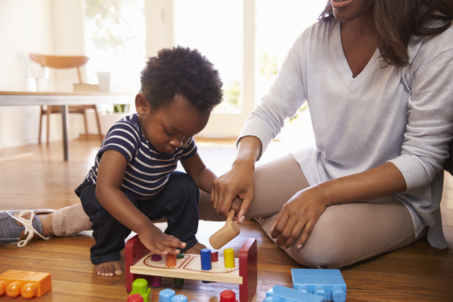 a 19 month old child plays with a developmental toy while crouching next to a female adult