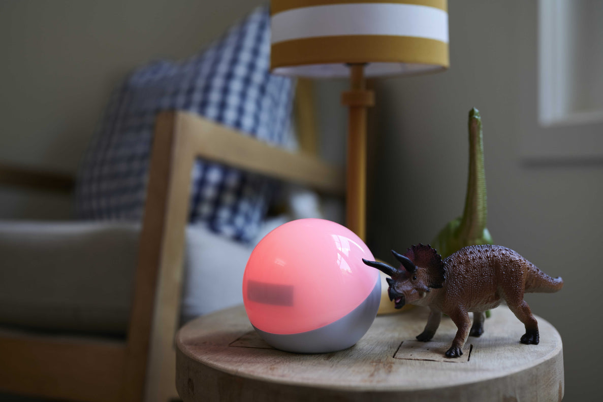 A glowing pink, spherical OK-to-Wake light sits on a wooden side table next to a toy Triceratops and a table lamp