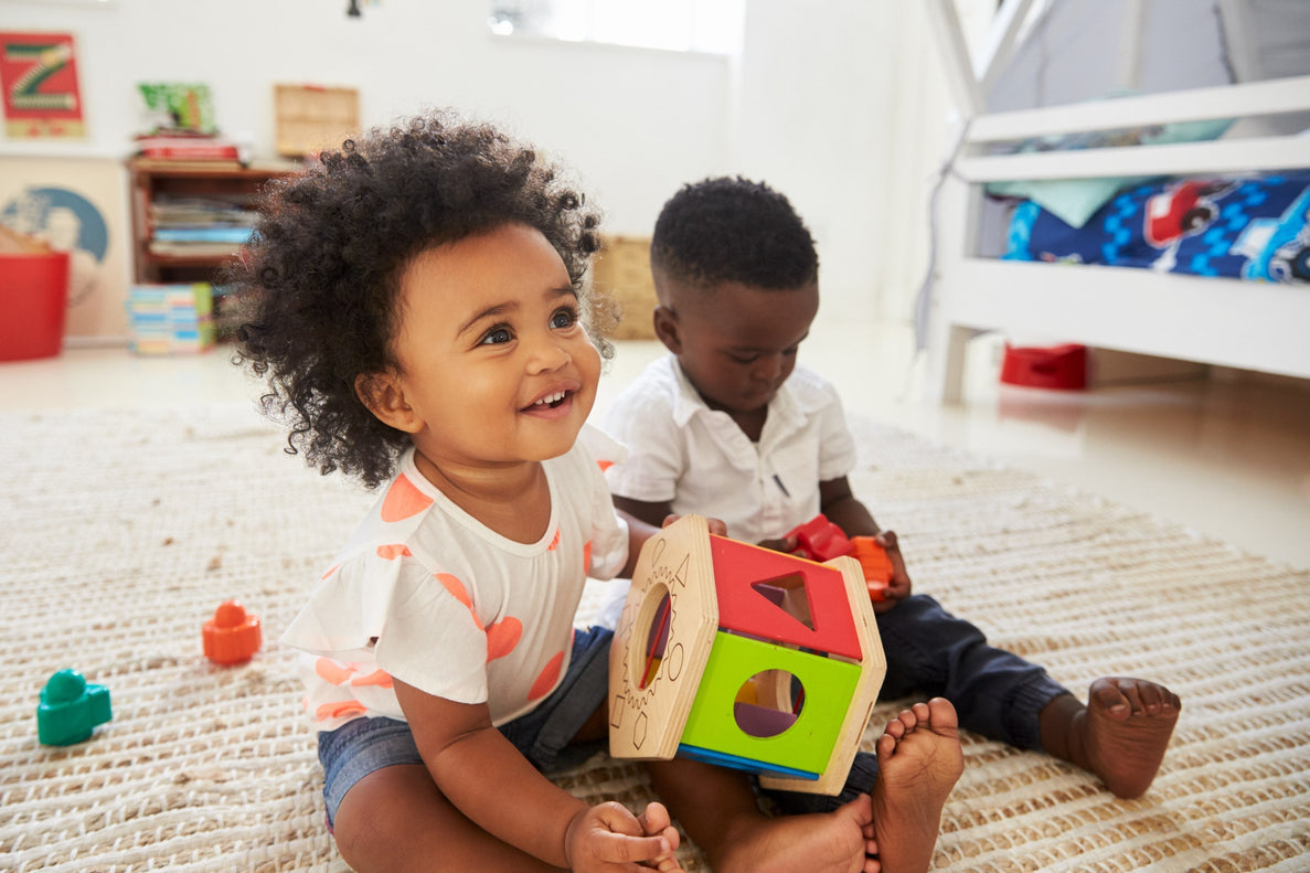 a 23 month old girl plays with a sorting toy while sitting next to another toddler boy on the floor