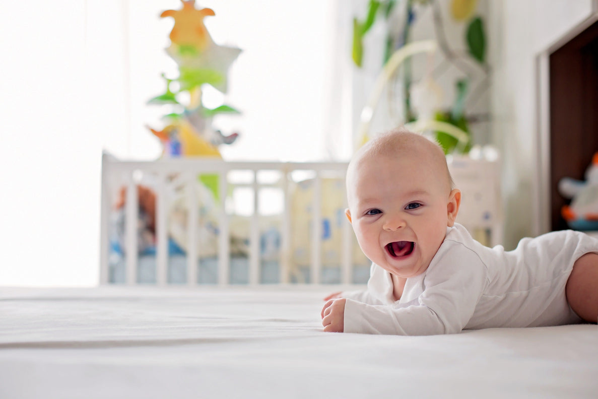 a joyful 4 month old baby in a white onesie crawls on a white rug with a crib in the background
