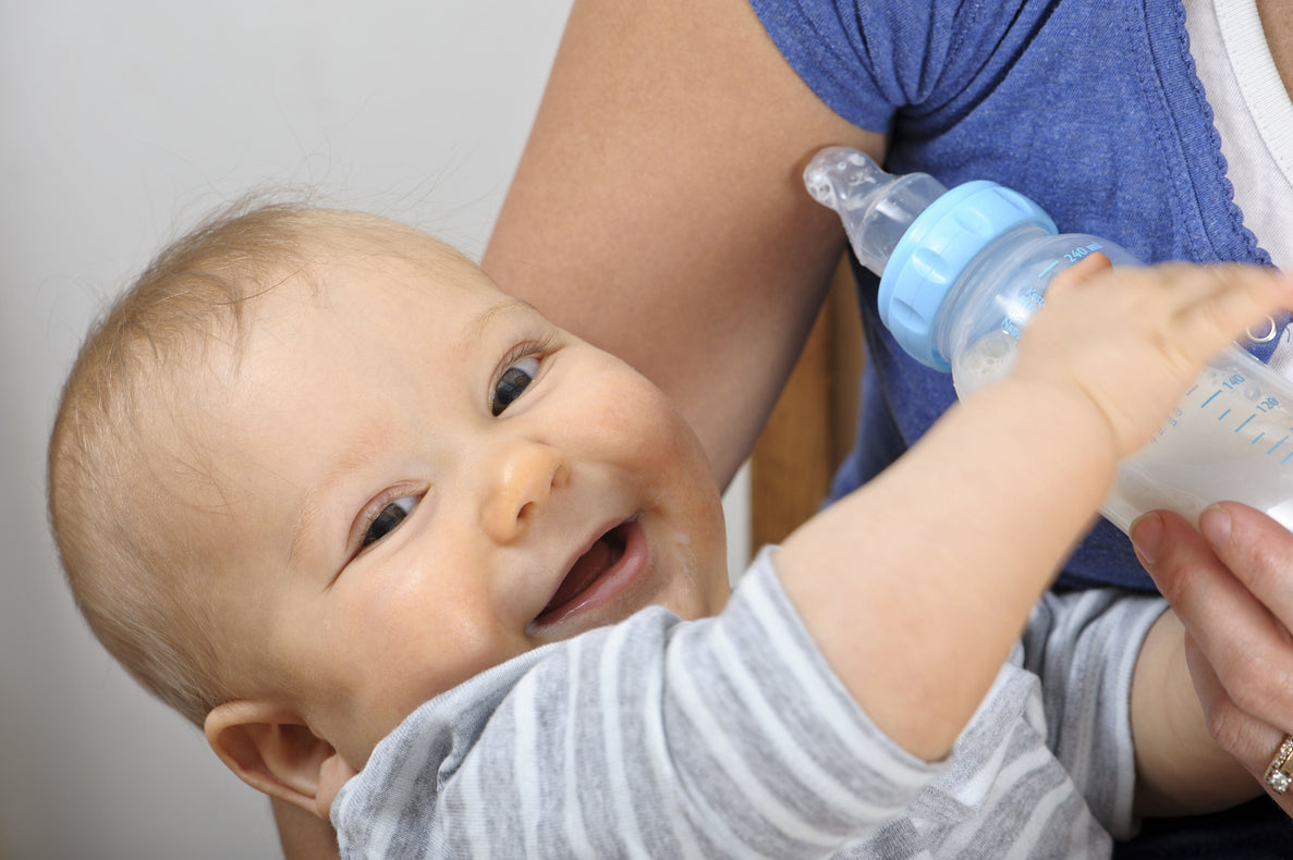 a smiling baby is held by an adult for a feeding while reaching for a bottle