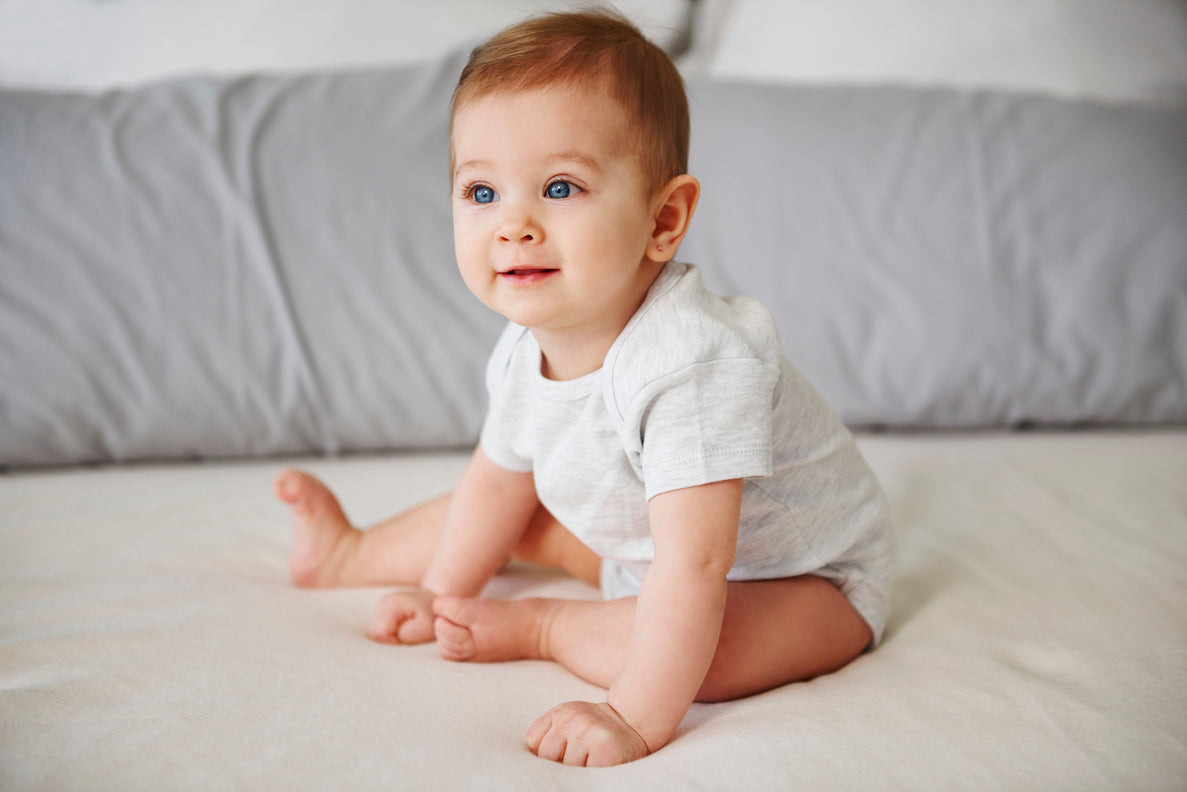 a 7 month old baby dressed in a white onesie practices sitting up on a bed with grey pillows in the background