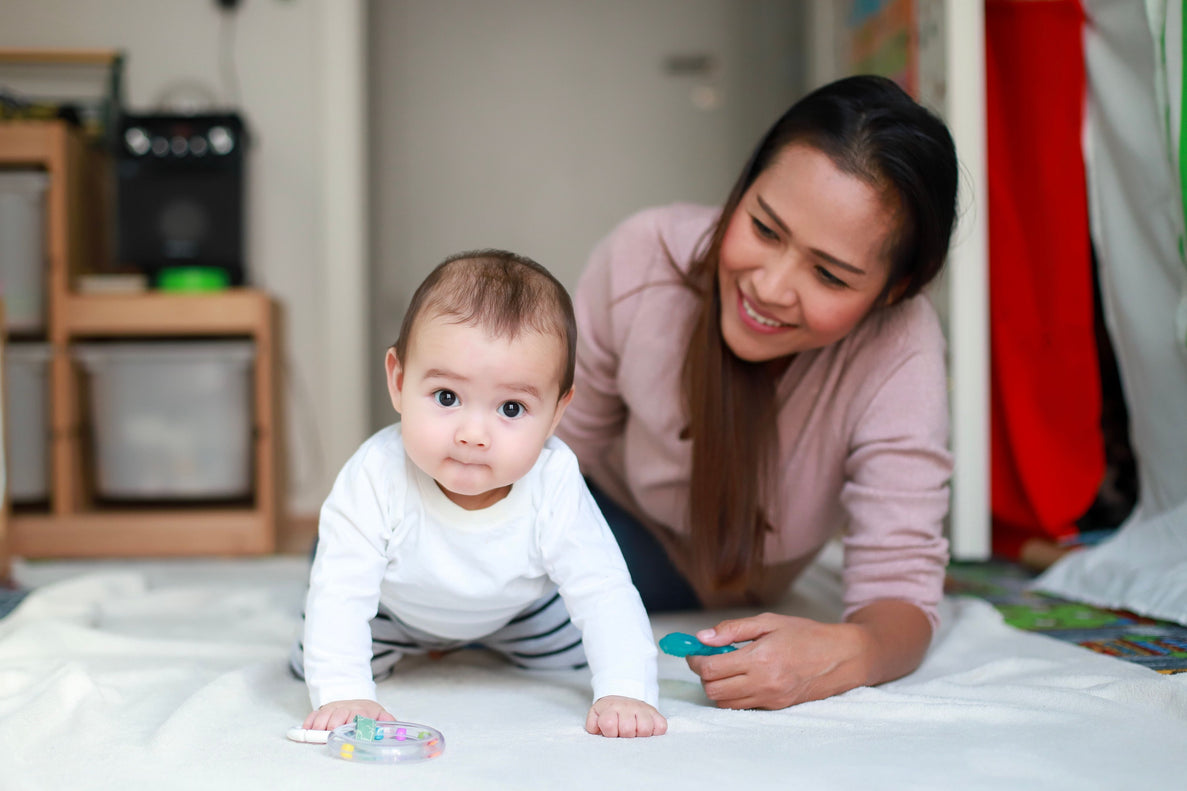 an 8 month old baby crawls towards a ring toy on a blanket with their mom next to them smiling and offering support