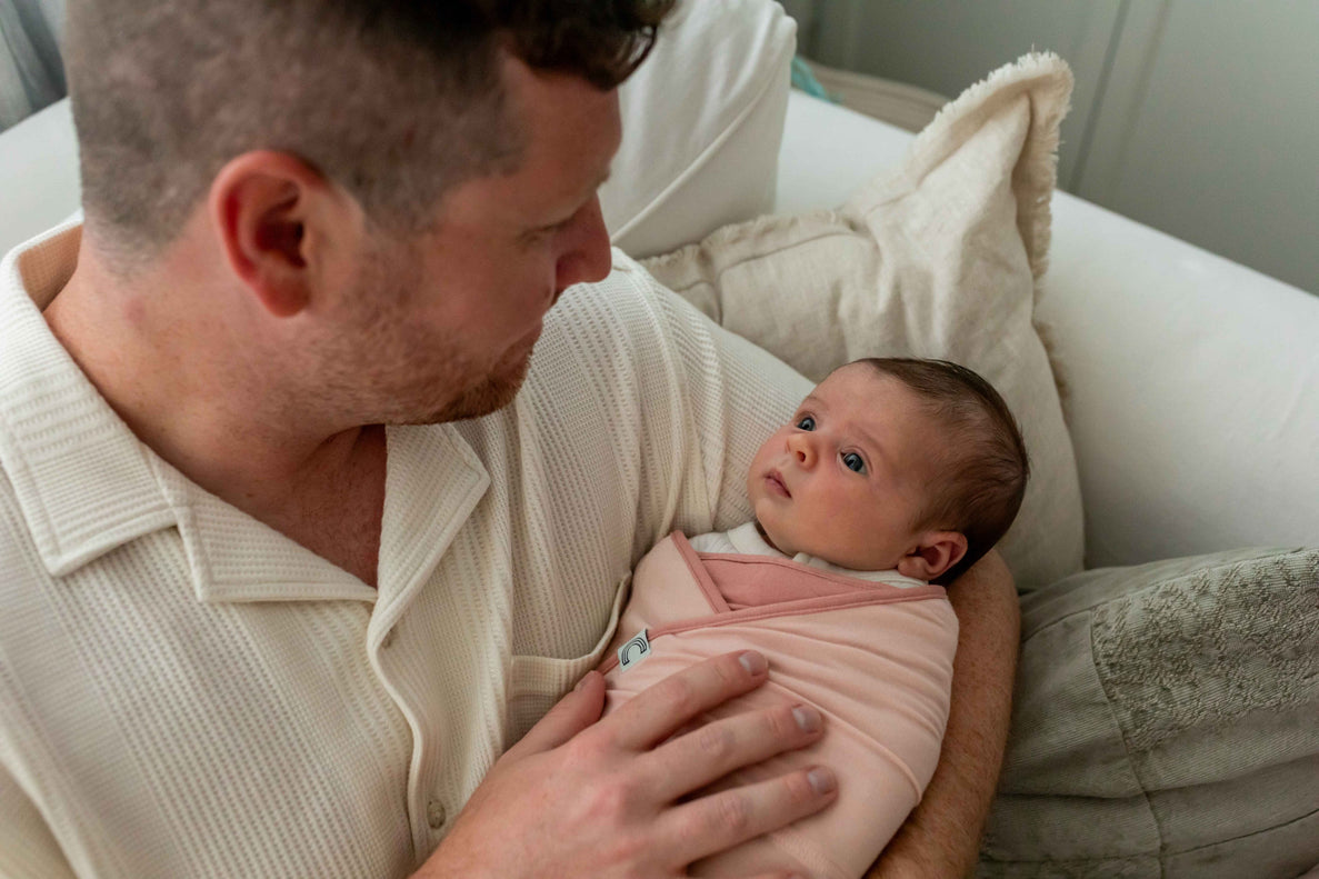 a dad holds his newborn wrapped in a pink b.e.s.t swaddle. The baby looks at him inquisitively