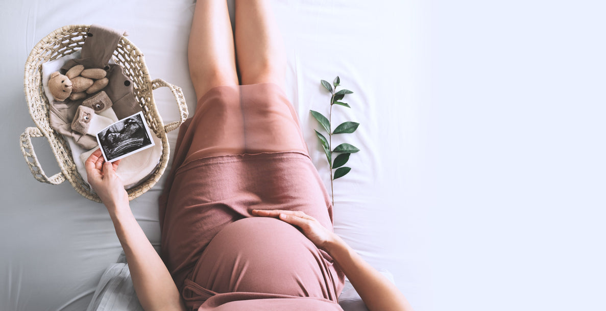 a waist down photo of a pregnant woman wearing a mauve dress touching her belly and holding a sonogram picture over a small basket full of baby items.