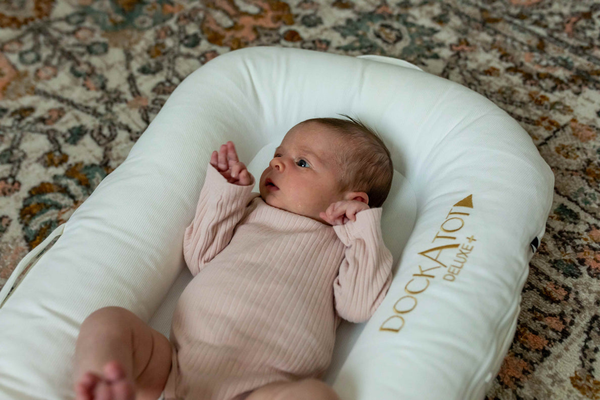 a newborn baby with a pink onesie looks at her hand while laying in a DockATot baby lounger