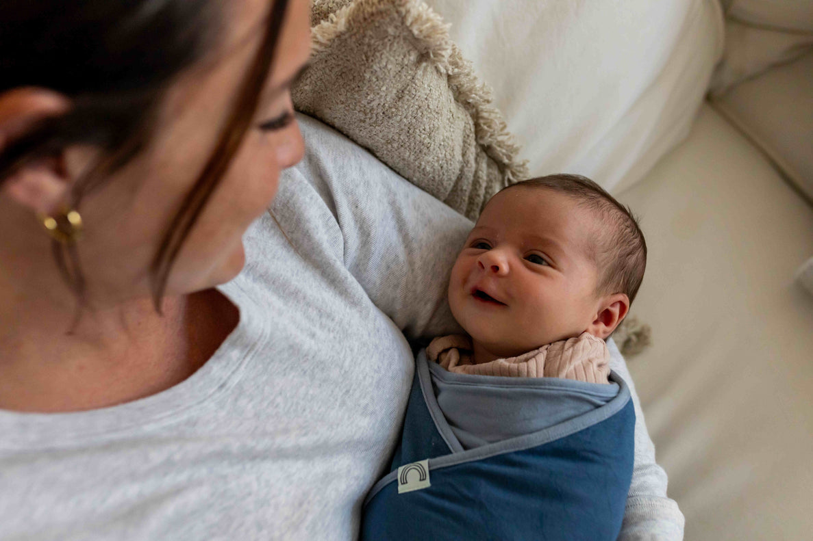 a newborn wrapped in a blue swaddle is smiling as they look a their mom who is holding them.