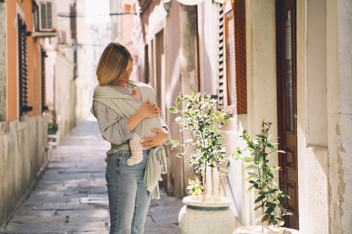 a mom is babywearing as she walks down the street. She cuddles the baby as they are sleeping on her
