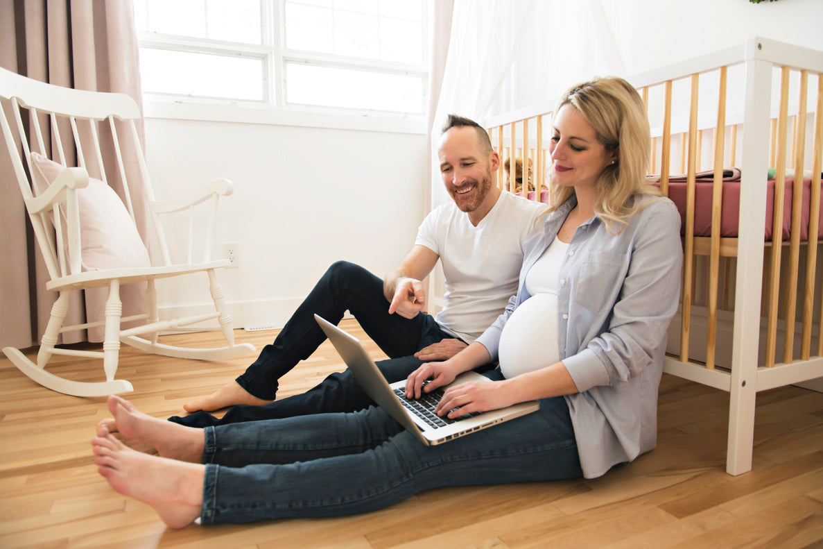 An expectant mom with a pregnant belly sits on the floor leaning against a crib typing on a laptop. Her male partner sits next to her.