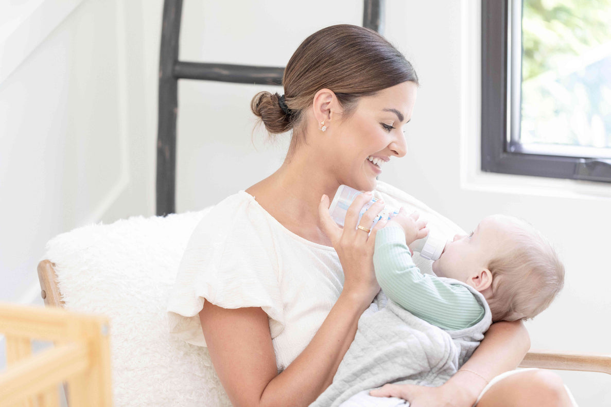 a mom smiles at her baby as she feeds the baby with a bottle while holding them in her arms