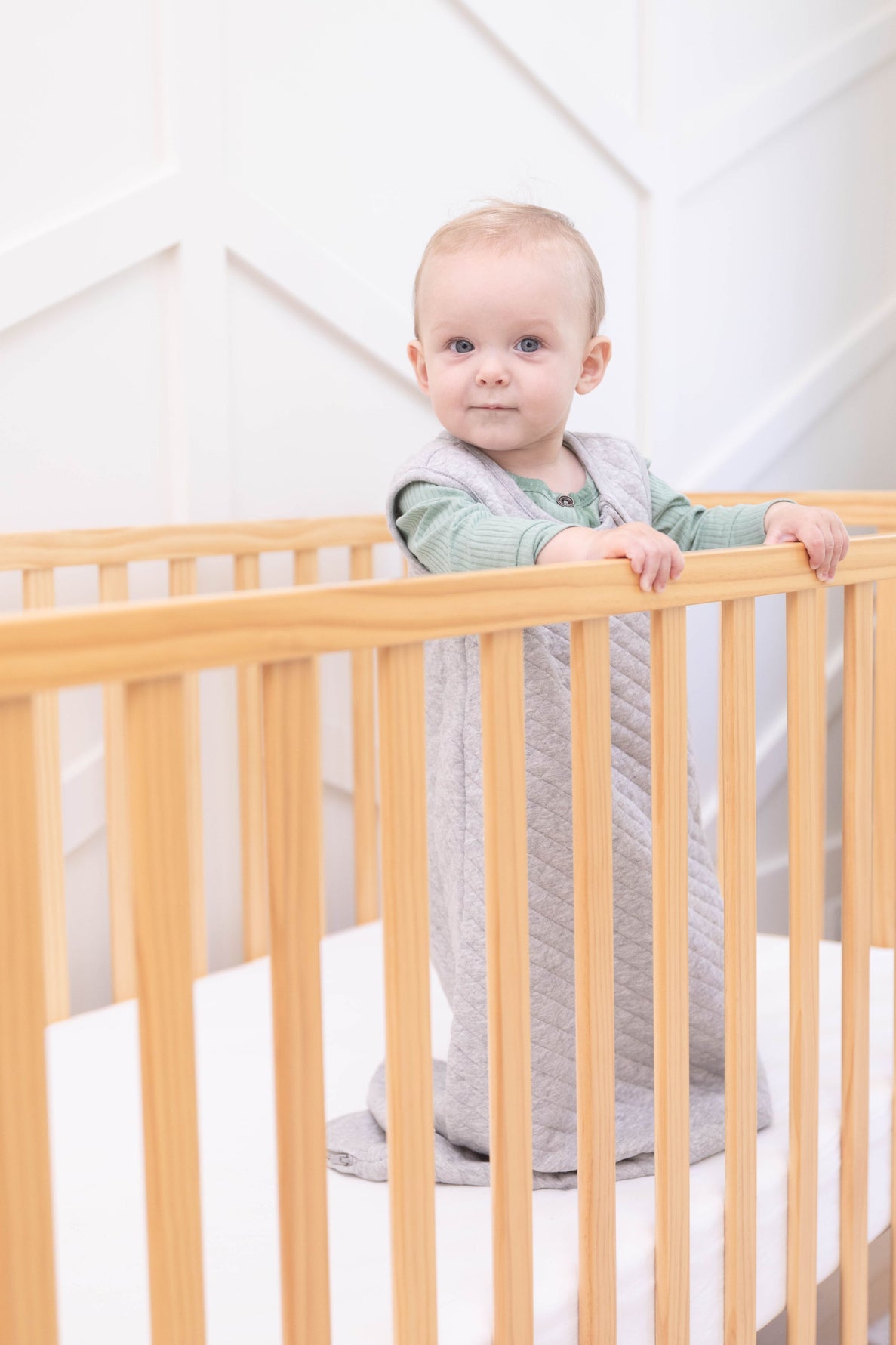 a content looking baby wearing a sleep sack stands in their crib holding on to the rail