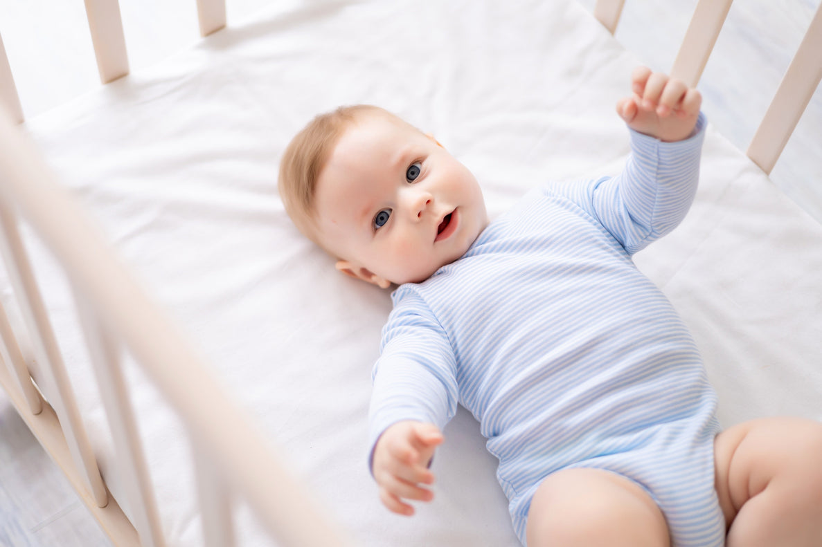 a content looking baby in a blue onesie lays in his crib with his arms raised up