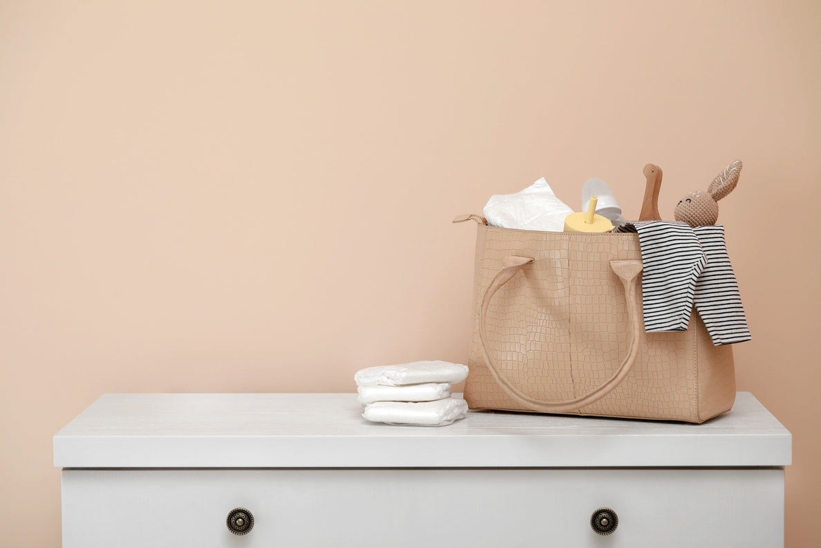 a photo of a diaper bag with baby items spilling out of the top. 3 diapers are stacked next to the bag sitting on top of a white dresser