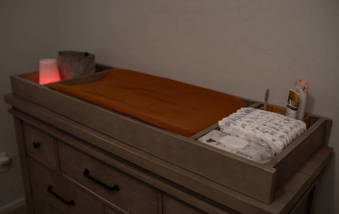 a diaper changing table setup in a dimly lit room. There is a red nightlight on the table along with diapers, wipes, and ointment. The changing pad has a rust colored cover.