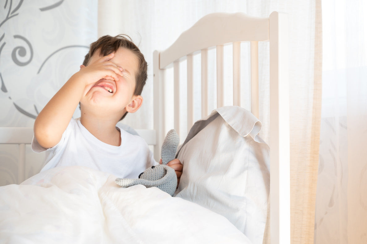 a toddler boy holds his head and looks like he;s upset or frustrated. He is holding a lovey in his hand and laying in his bed