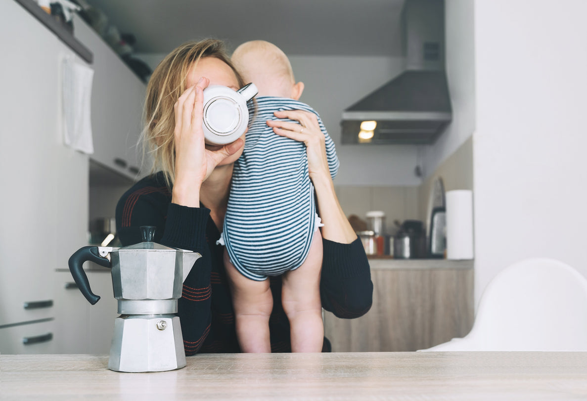 A tired mom chugs coffee while holding a small baby over her shoulder. A coffee pot sits on the table in front of her with a kitchen in the background