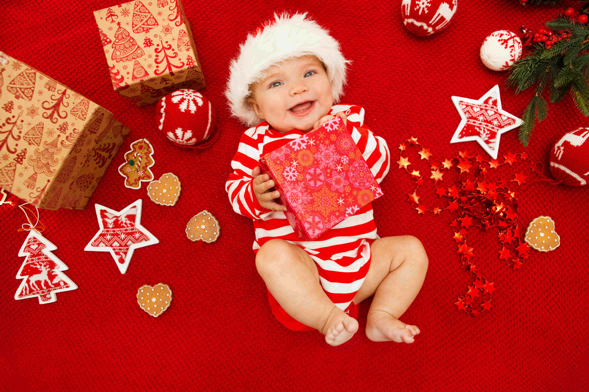 A baby in a red and white striped onesie and a Santa hat lies on a bright red blanket surrounded by holiday decor.