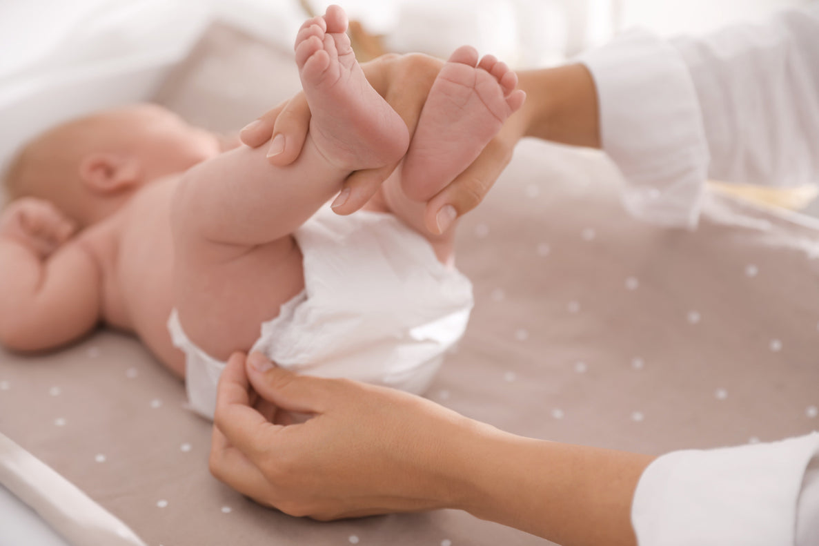 A close-up image shows a person's hands changing a baby's diaper on a changing table or mat. The baby is lying on its back with its legs held up by one hand, exposing the white disposable diaper being fastened by the other hand.