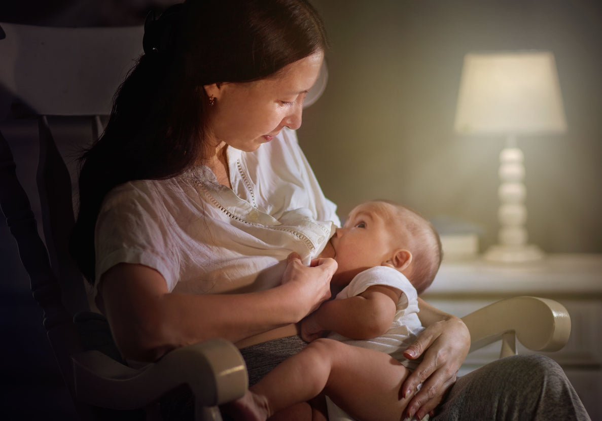 a mom nurses her baby while sitting in a darkened room with a lamp lit in the background