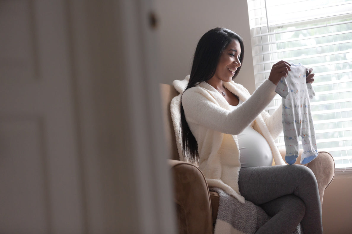 a pregnant mom is sitting in a chair in the nursery folding baby clothes with a content smile