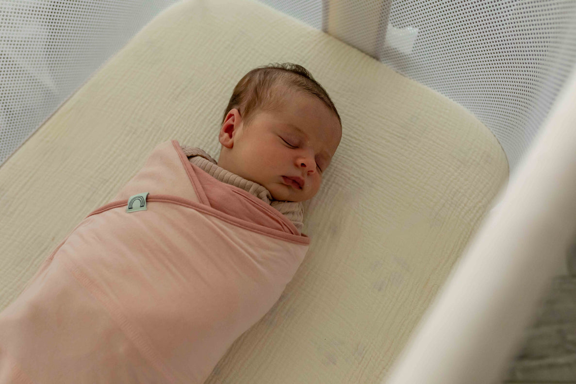 a sleeping newborn wrapped in a pink b.e.s.t. swaddle lays on a cream colored bassinet mattress