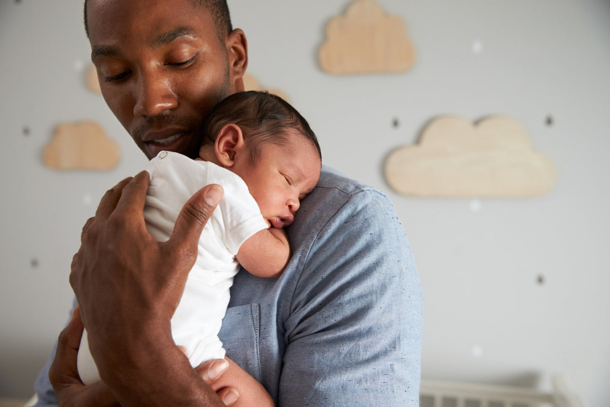 A dad holds his sleeping newborn over his shoulder. There is a nursery background with clouds hanging on the wall