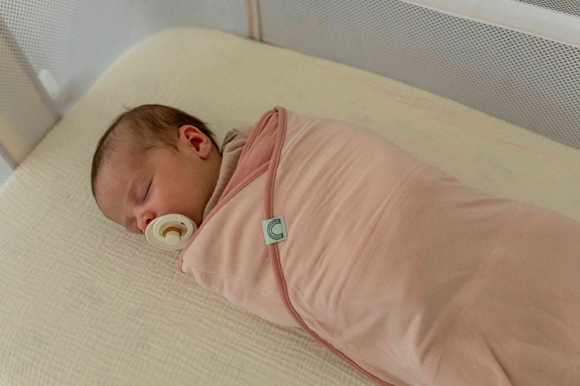 an image of a sleeping newborn baby in a bassinet. The baby is swaddled in a pink swaddle with a pacifier in their mouth
