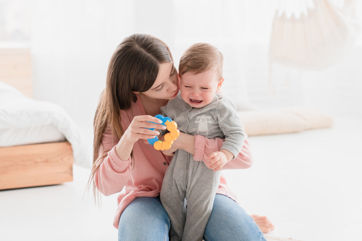 A mom sits on the floor holding her baby in one hand and a toy in the other hand. The baby is crying and mom is trying to comfort him with the toy and affection.