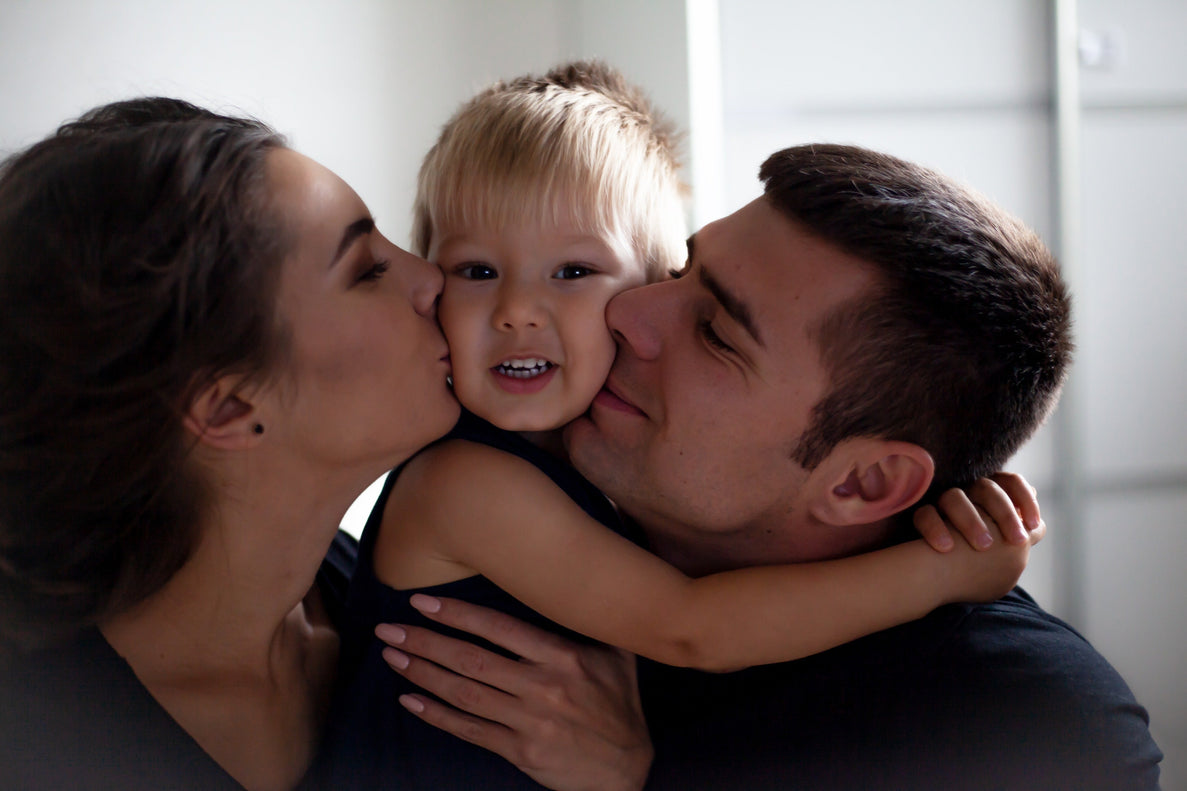 a toddler is sandwiched between his mom and dad as they hug and kiss him affectionately