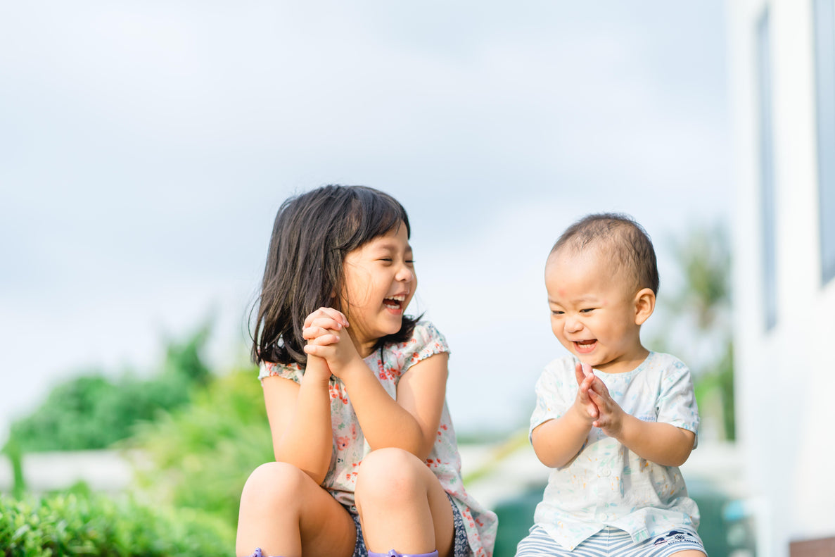 A young girl and a baby are sitting side-by-side outdoors, both laughing. The girl, with long dark hair, is looking at the baby and clapping her hands together. The baby is also clapping.