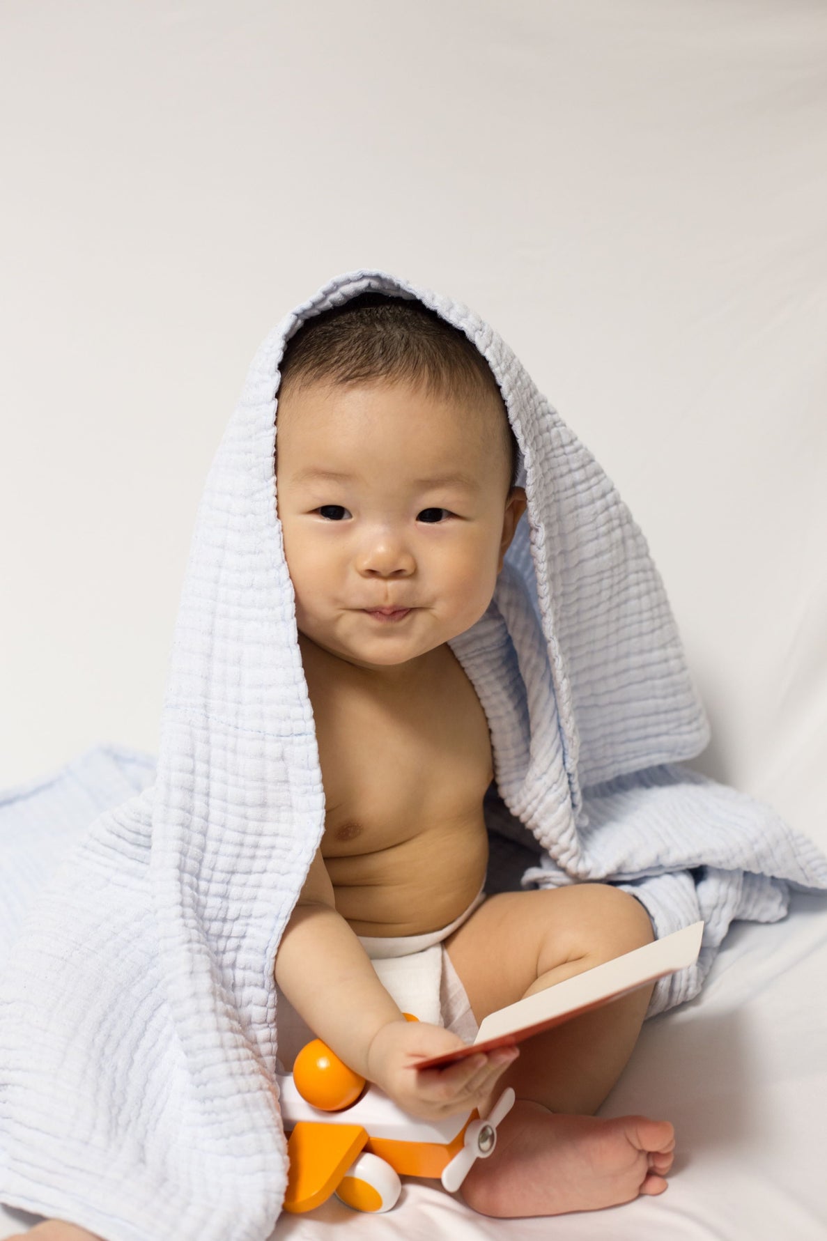 A smiling 6 month old baby sits wrapped in a light blue blanket, holding an orange toy airplane and a small book. The baby is wearing a diaper.