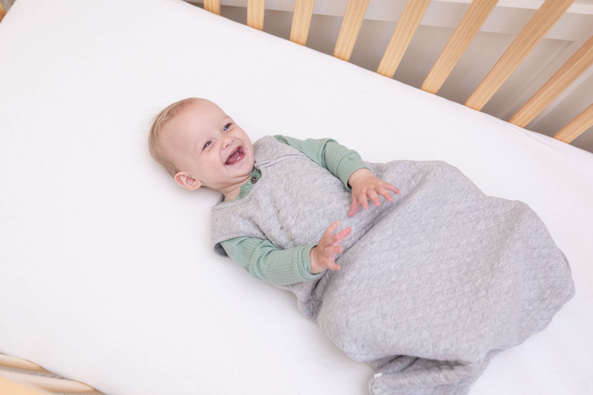 a baby laughs playfully while laying in his crib wearing a grey sleep sack