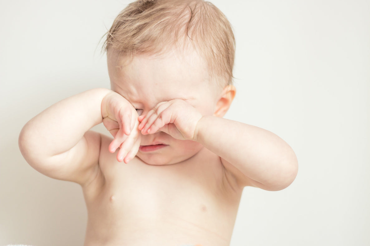 A baby with no shirt on rubs his eyes sleepily
