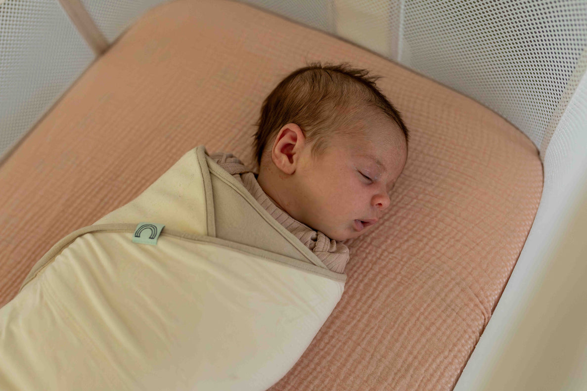a newborn baby is sleeping with their head turned to the side wrapped in a cream colored swaddle.