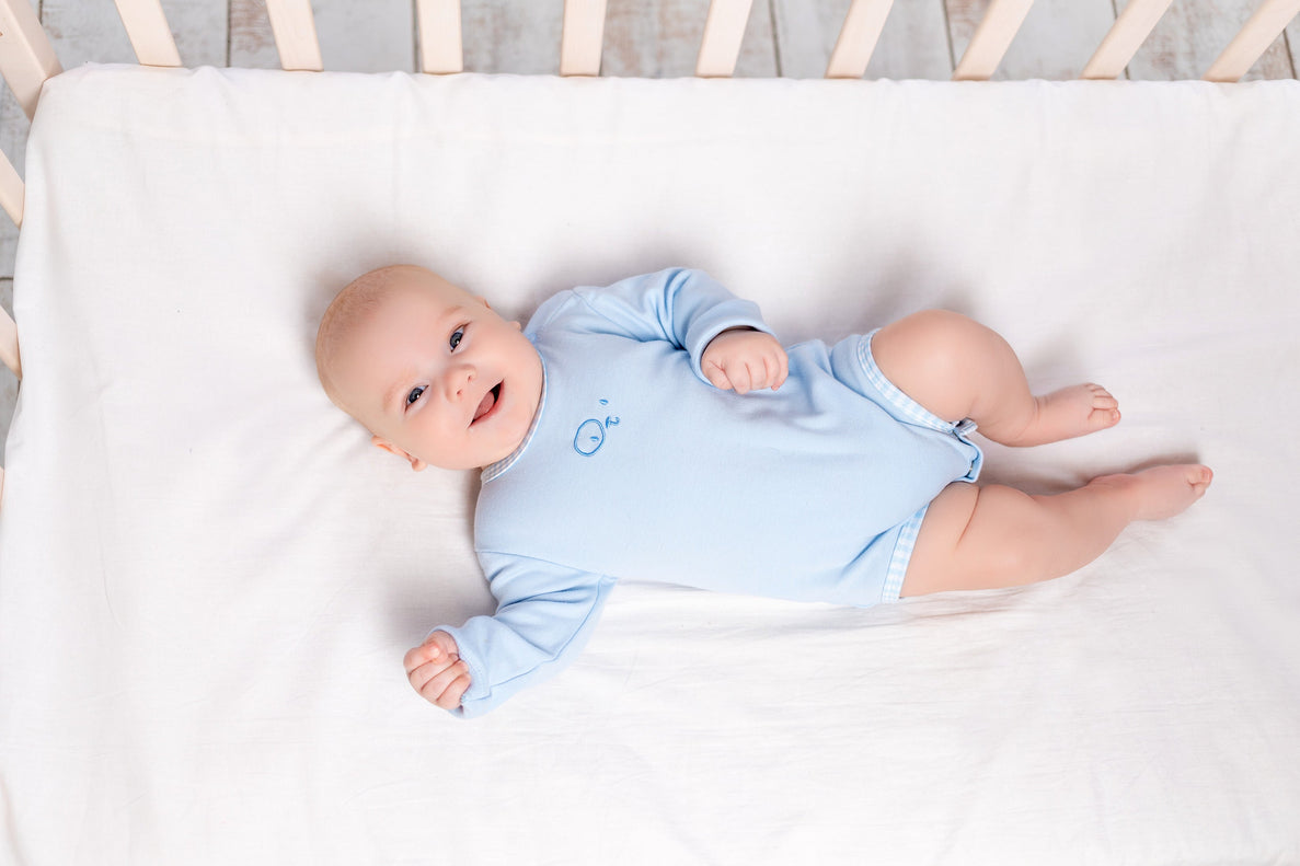 A smiling baby lays in a crib with a white sheet smiling. He wears a baby blue onesie