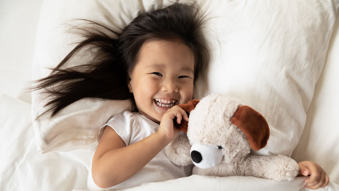 a toddler girl smiles joyfully as she lays in bed with a stuffed dog next to her