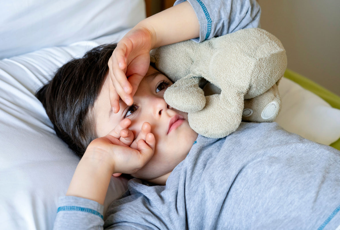 A toddler lying in bed looks unhappy as he covers his face with his hands and a stuffed animal.