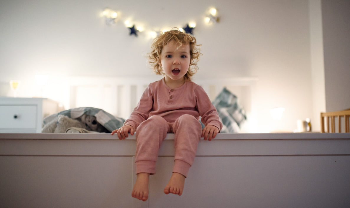 A toddler looks mischievous as he sits on the edge of his bed.