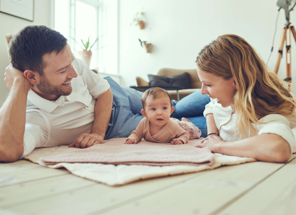 A mom, dad, and newborn all lay on the floor smiling. The baby is practicing tummy time on a blanket as parents observe them proudly
