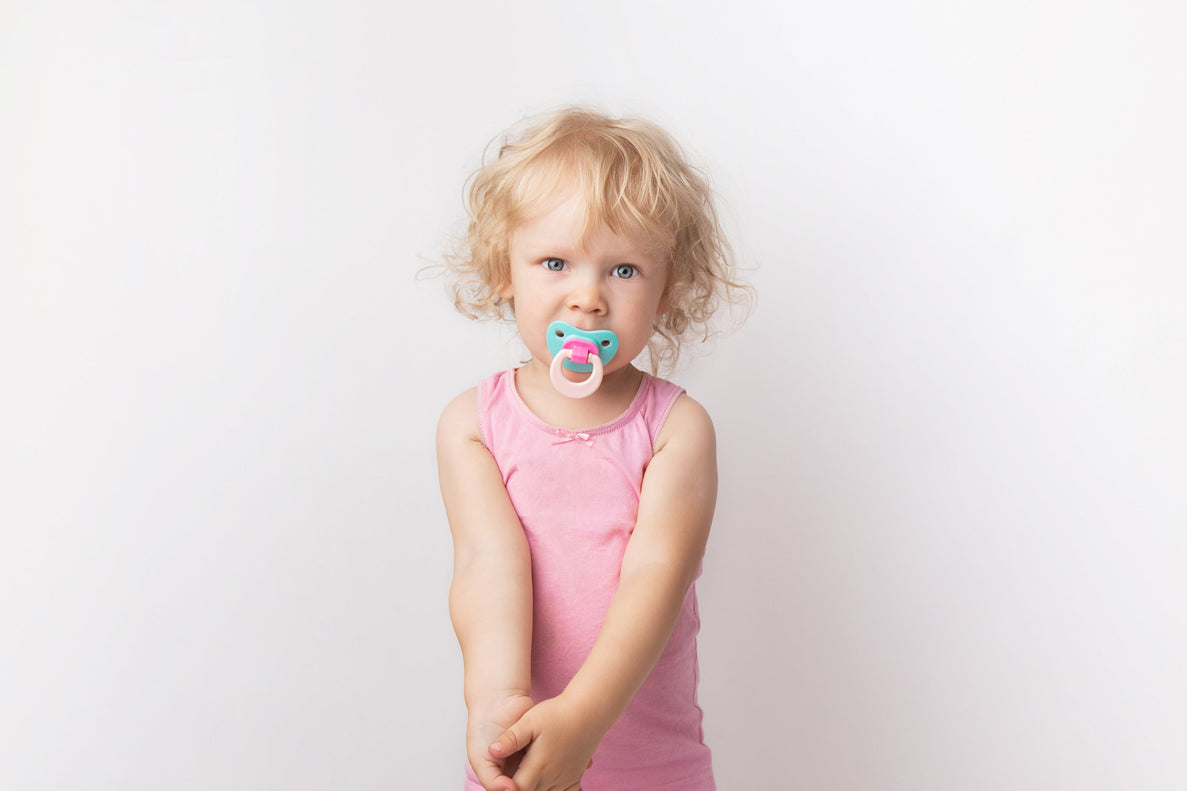 a toddler stands against a white background with a blue pacifier in her mouth