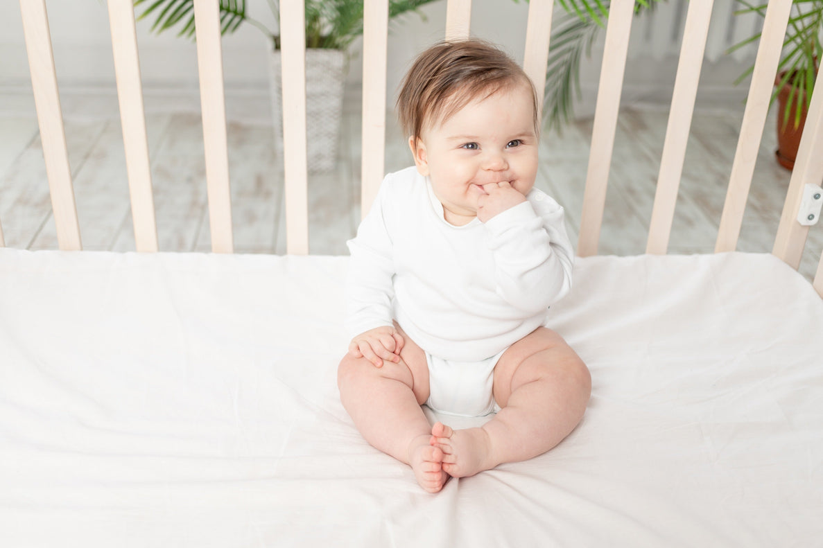 a baby sits up in a crib with his hand in his mouth. He looks happy