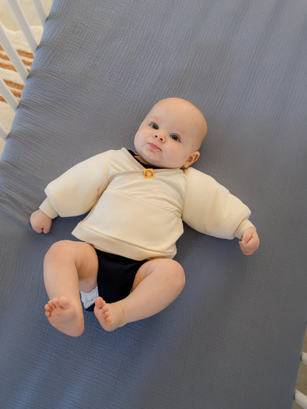 baby wearing coconut cream Jacky swaddle transition jacket layered over black onesie laying on back in a crib