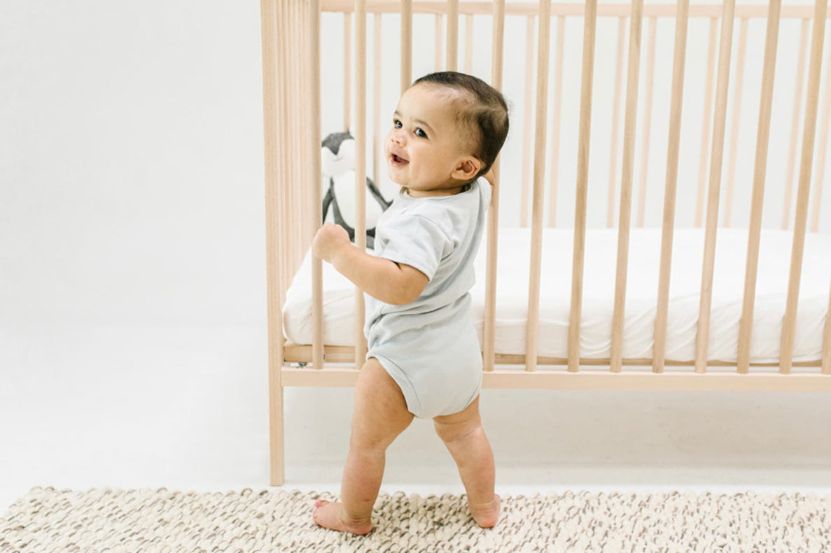 a 12 month old baby stands while holding his crib with one hand looking proud