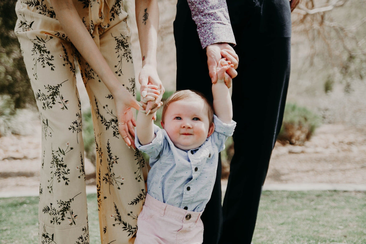 a 14 month old baby smiles as they try to walk holding on to their parents hands