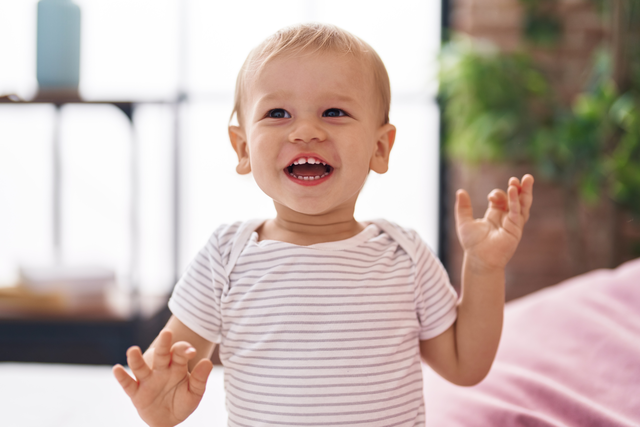 a joyful 15 month old baby holds their hands up and smiles