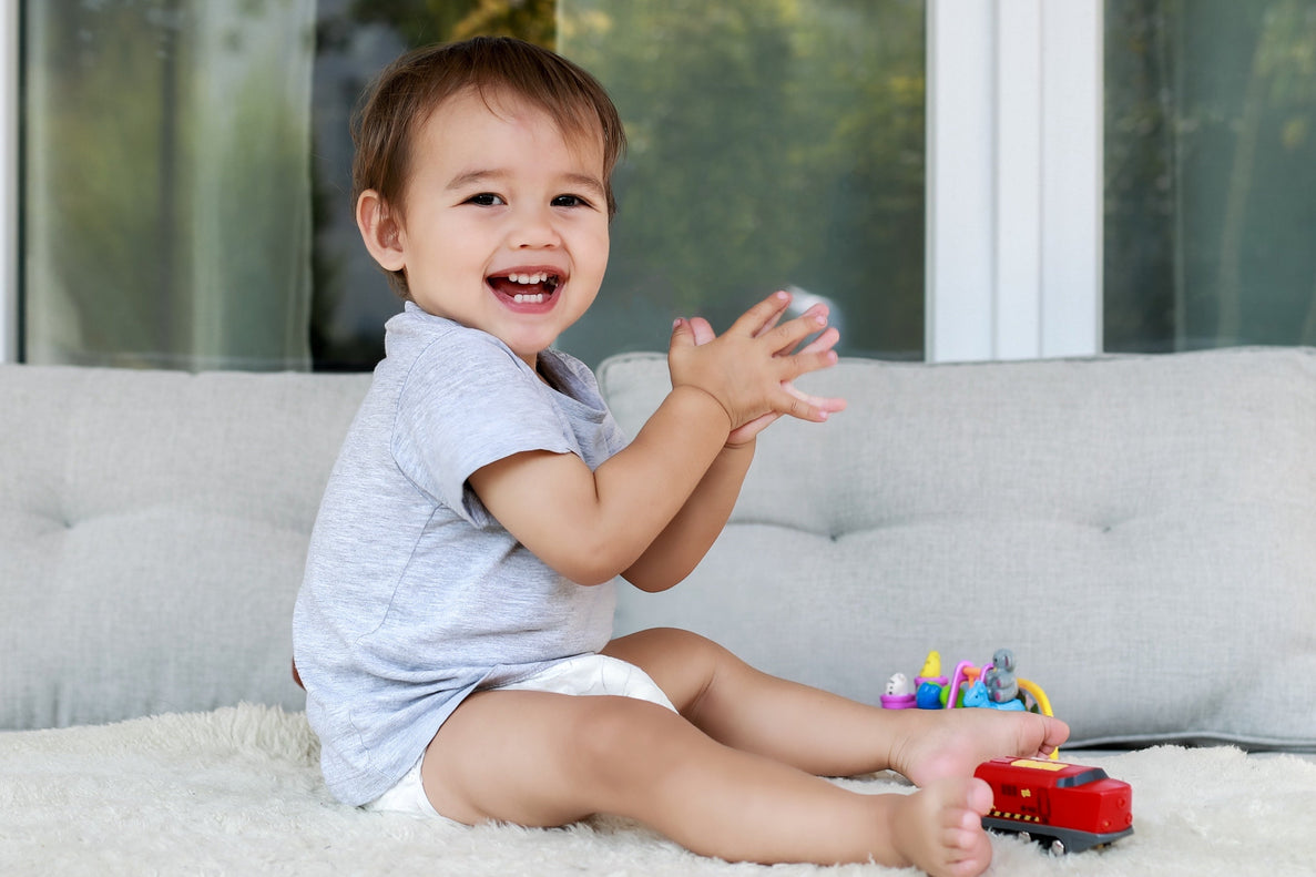 a 16 month old baby dressed in a tshirt and diaper claps with toys scattered at his feet