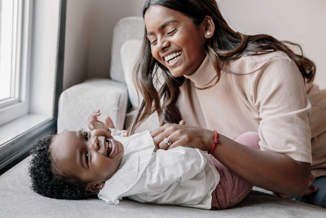 a joyful mom and 17 month old baby laugh while mom leans over the baby tickling them