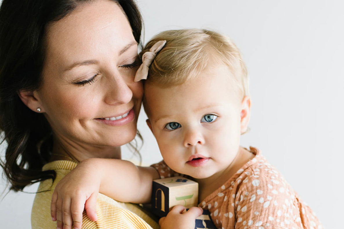 an 18 month old baby looks seriously at the camera while her mom holds her and looks at her lovingly