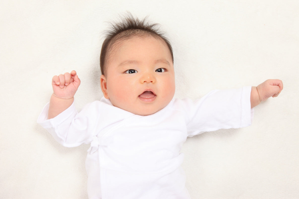 A happy two-month-old baby with dark hair, lying on their back in a white onesie, smiling and raising their hands